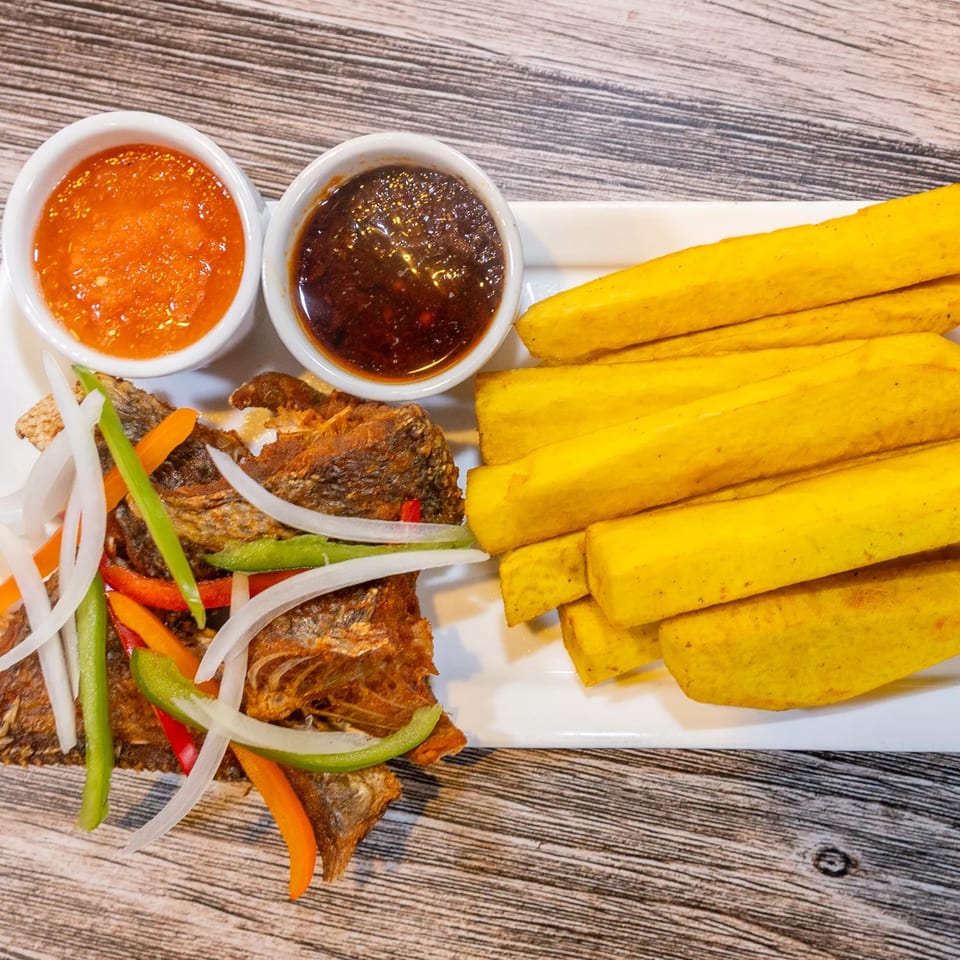 Fried fish with sliced onions, peppers, plantain fries, and two dipping sauces on a white plate.