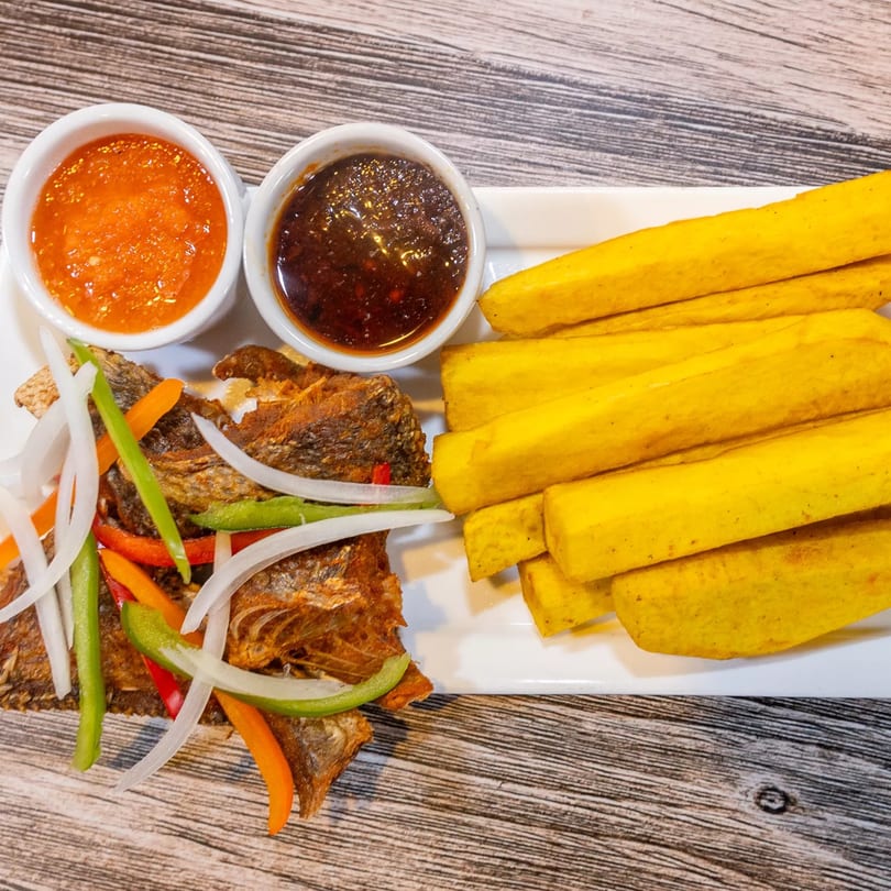 Fried fish with sliced onions, peppers, plantain fries, and two dipping sauces on a white plate.
